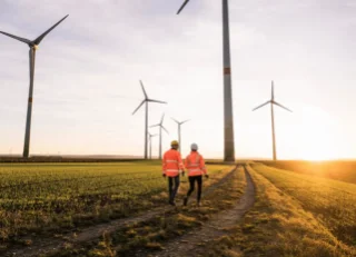 Two people in safety gear walk on a dirt path through a wind farm at sunset. Tall wind turbines surround them, casting long shadows on green fields.