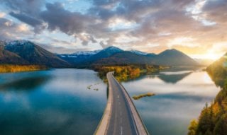A paved road crosses over a calm lake, surrounded by mountains