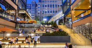 Modern mixed-use plaza with outdoor seating, shops, and evening lighting.
