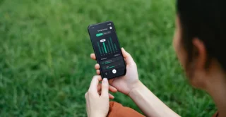A person holding a smartphone in a grassy area, displaying a dark-themed app with green graphs indicating energy usage. The scene feels calm and focused.