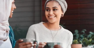 Two women sit together, smiling while holding mugs. One wears a light sweater and headscarf; the other a hijab. The scene is warm and friendly.