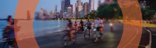Blurry cityscape at dusk with cyclists riding along a waterfront path.