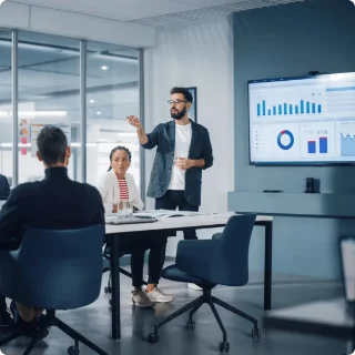 A man presents data on a screen to colleagues in an office meeting room. Charts and graphs are displayed, conveying a professional and collaborative atmosphere.