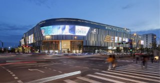 Modern shopping mall exterior with illuminated facade and busy city street at dusk.