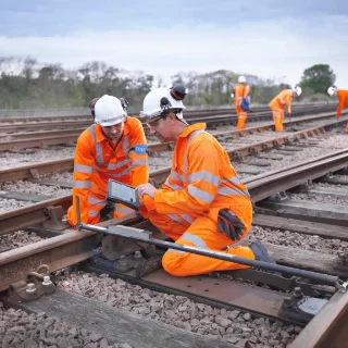 Railway workers in orange safety gear inspect tracks, using a tablet, surrounded by gravel and rails. Overcast sky. Teamwork and focus are evident.