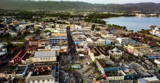 Aerial view of a vibrant coastal cityscape with colorful buildings, bustling streets, and a central roundabout. Mountains and calm ocean in the background.