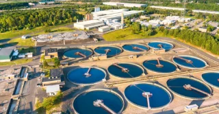 Aerial view of a wastewater treatment plant with multiple circular sedimentation tanks, surrounding buildings, and green landscape.
