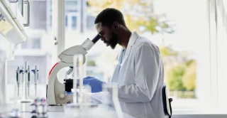 A scientist wearing a white lab coat and blue gloves examines a sample through a microscope in a bright laboratory, indicating focus and precision.