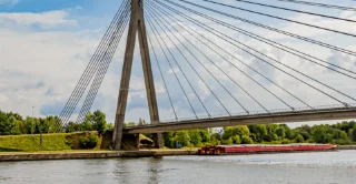 A modern suspension bridge with numerous cables spans a calm river. Below, a red barge sails smoothly. Green trees and a partly cloudy sky are in the background.