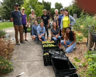 A group of people smiles near crates filled with harvested green fruits in a lush community garden. The setting is relaxed and collaborative.
