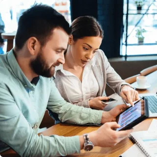 A man and woman work together in a bright café, focused on a tablet. She gestures at the screen, while he holds it. Open laptop and coffee nearby.