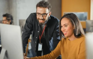 A man and woman smile as they look at a computer screen in a modern office. The man wears glasses and a lanyard. The atmosphere is collaborative.
