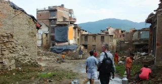 A group of people walks through a street lined with damaged brick buildings. Debris is scattered, and mountains are visible in the background.