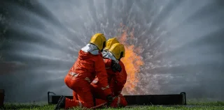 Three firefighters in orange suits and yellow helmets kneel while battling a blaze.