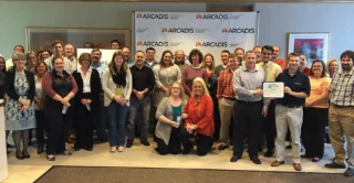 A group of 30 people, smiling in a room, pose for a photo. Some hold a certificate. The backdrop features multiple "Arcadis" logos. Casual office setting.