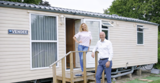 A man and woman stand smiling outside a beige mobile home with a "Vendee" sign. Wooden steps lead to the door. The setting is bright and cheerful.