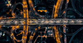Aerial night time view of a large highway interchange with multiple looping overpasses and moving traffic illuminated by bright city lights.