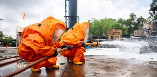 Two individuals in bright orange hazmat suits spray foam from a hose in an industrial setting, conveying urgency.
