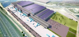 Aerial view of a large, modern warehouse complex with solar panels on the roof, surrounded by trucks, parking lots, and green spaces under a clear sky.