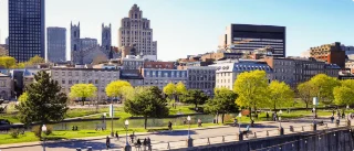 Urban park scene with people strolling along the pathway, surrounded by lush green trees. Historic buildings and a sunny skyline fill the background.