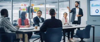 A diverse group of six people in a modern office engages in a meeting. A man stands presenting near a screen displaying graphs.