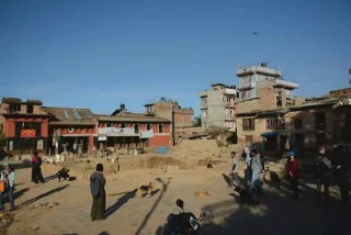 A busy urban square with people walking amongst old, rustic buildings and dogs lying on the ground under a clear blue sky.