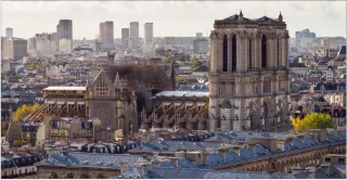 Aerial view of Notre-Dame Cathedral in Paris. Rooftops and modern city buildings are visible in the background.
