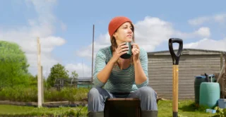 A woman in a red beanie, striped shirt, and boots sits on a crate, holding a mug in a garden. She looks pensive and relaxed under a clear sky.
